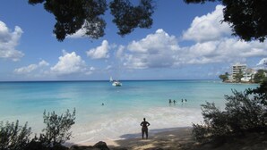Une plage à l’eau turquoise et limpide, une personne debout sur le rivage et un bateau au loin.