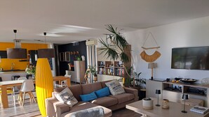A modern living room with a sofa, coffee table, and a TV. There is a kitchen area in the background with a yellow wall and a dining table.