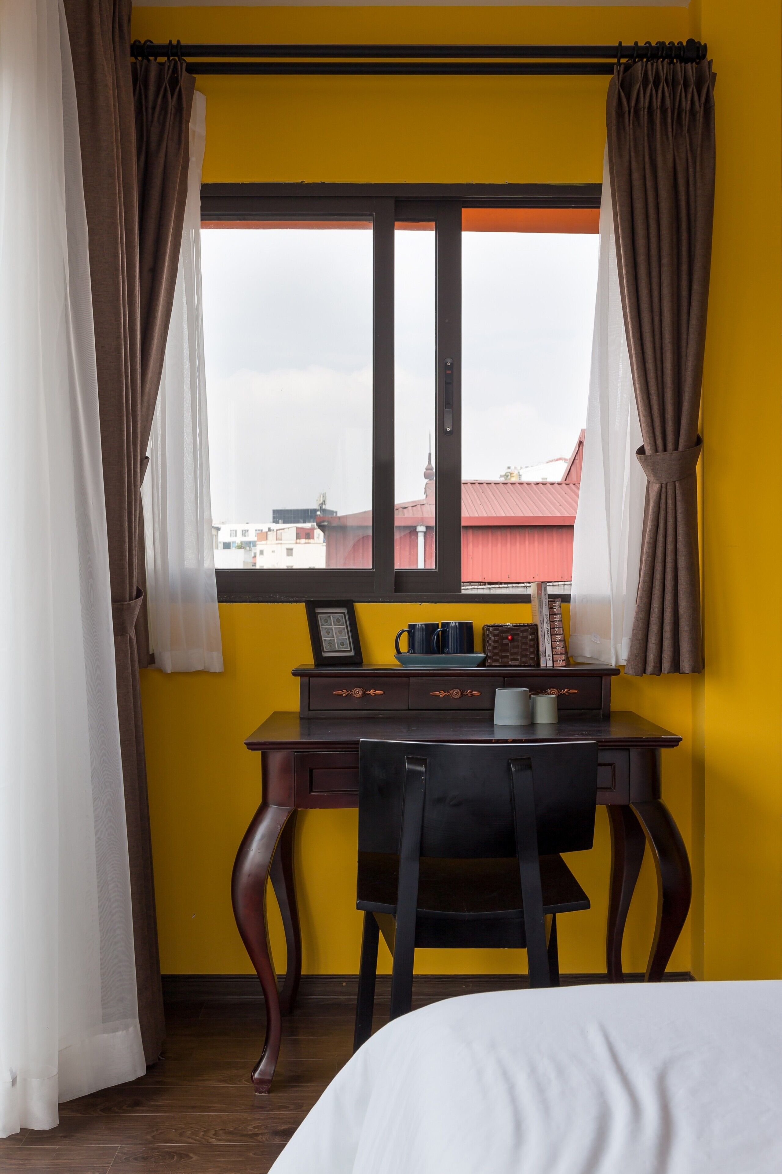A room with a yellow wall, a window with curtains, a desk with a chair, and a bed.