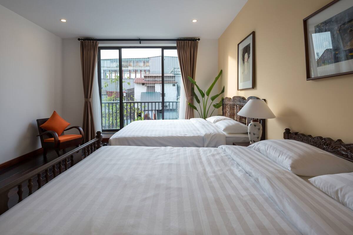 Quarto de hotel com duas camas, uma cadeira, um abajur e uma janela grande com cortinas.