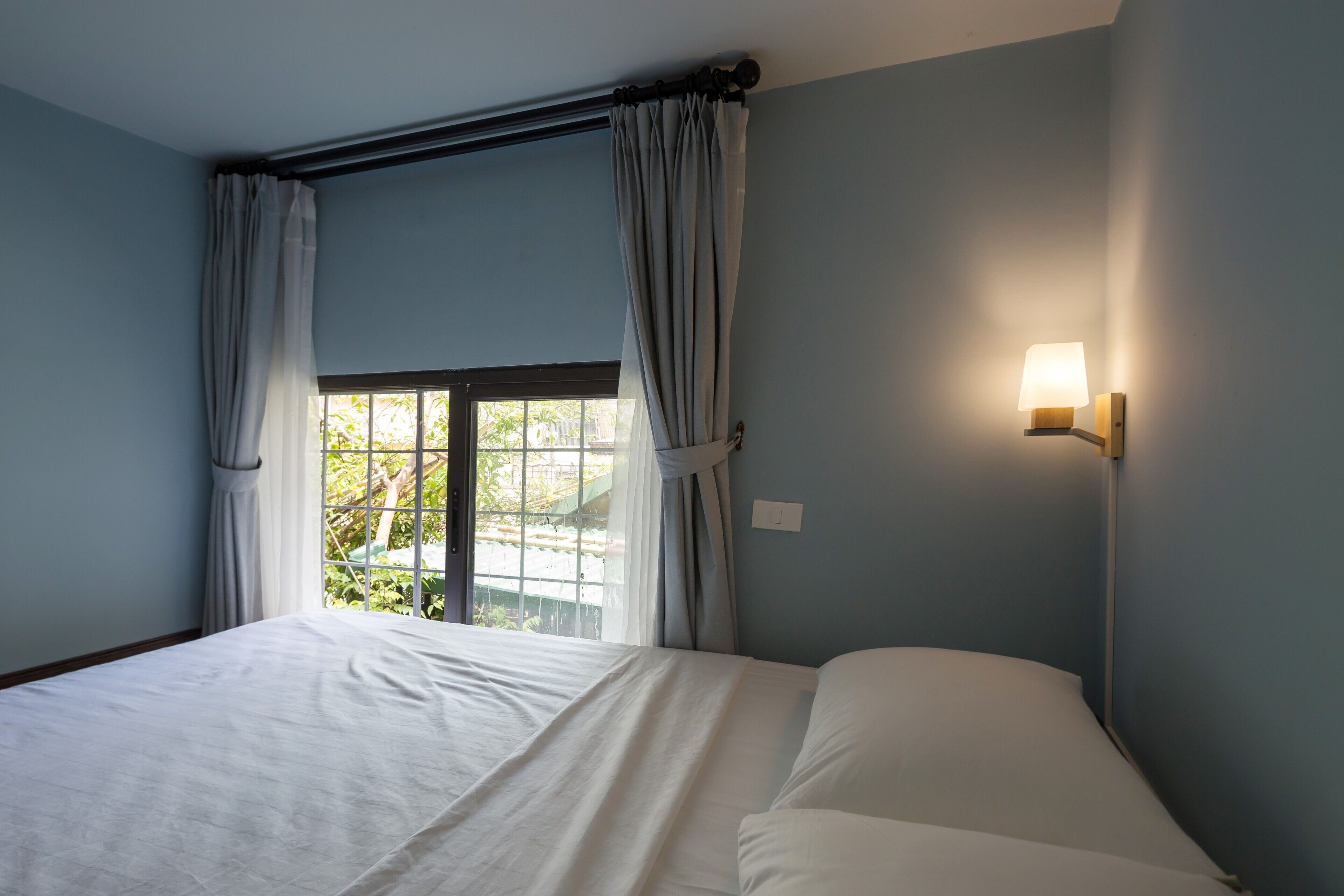 Un dormitorio con cama, ventana con cortinas y una lámpara fijada a la pared.