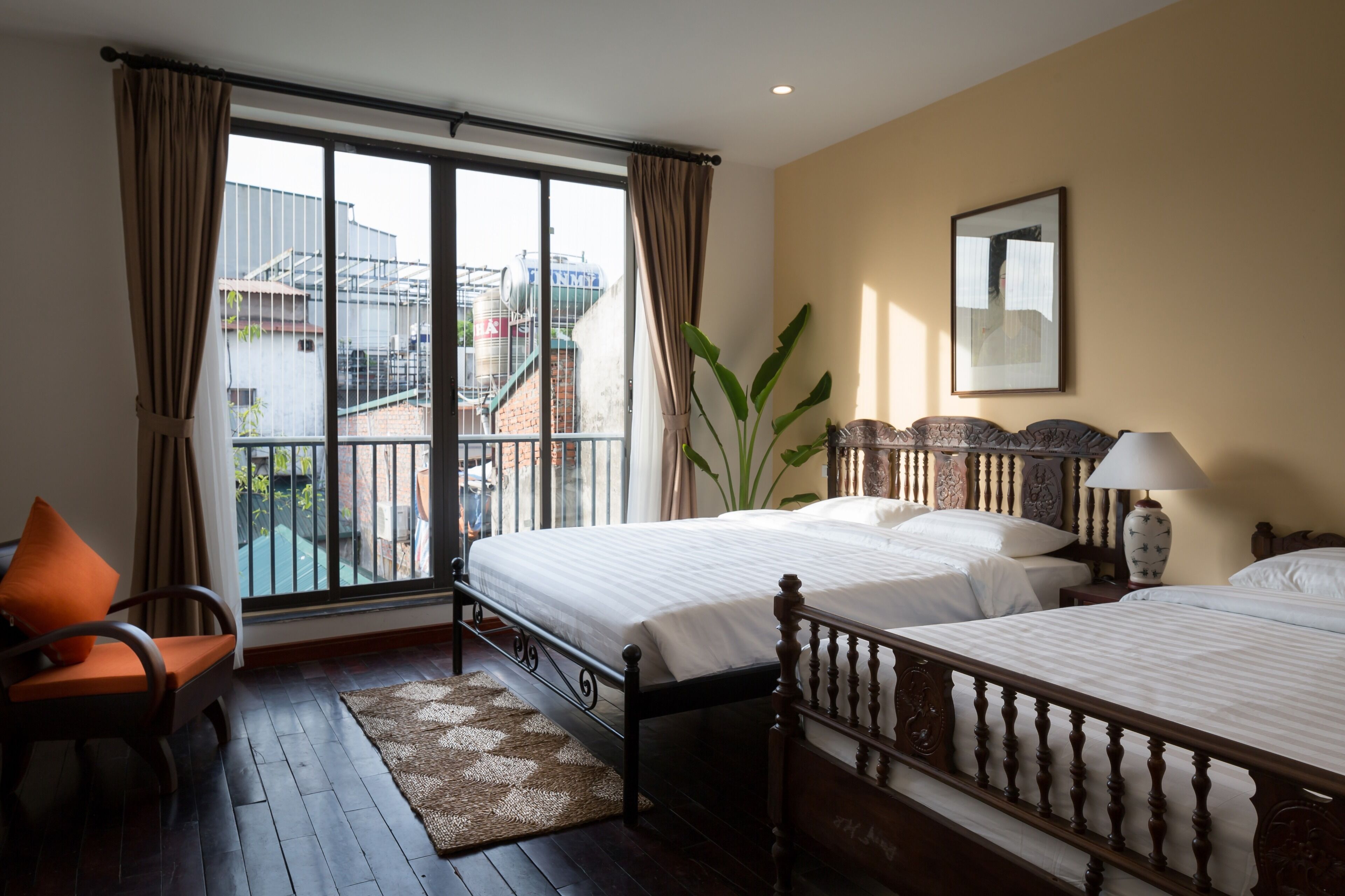 A hotel room with two beds, a large window with curtains, a view of buildings outside, a chair, and a lamp.