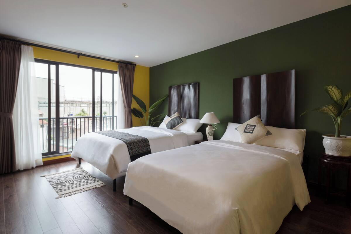 Quarto de hotel com duas camas, uma janela grande com cortinas, uma planta em vaso e vista para o exterior.