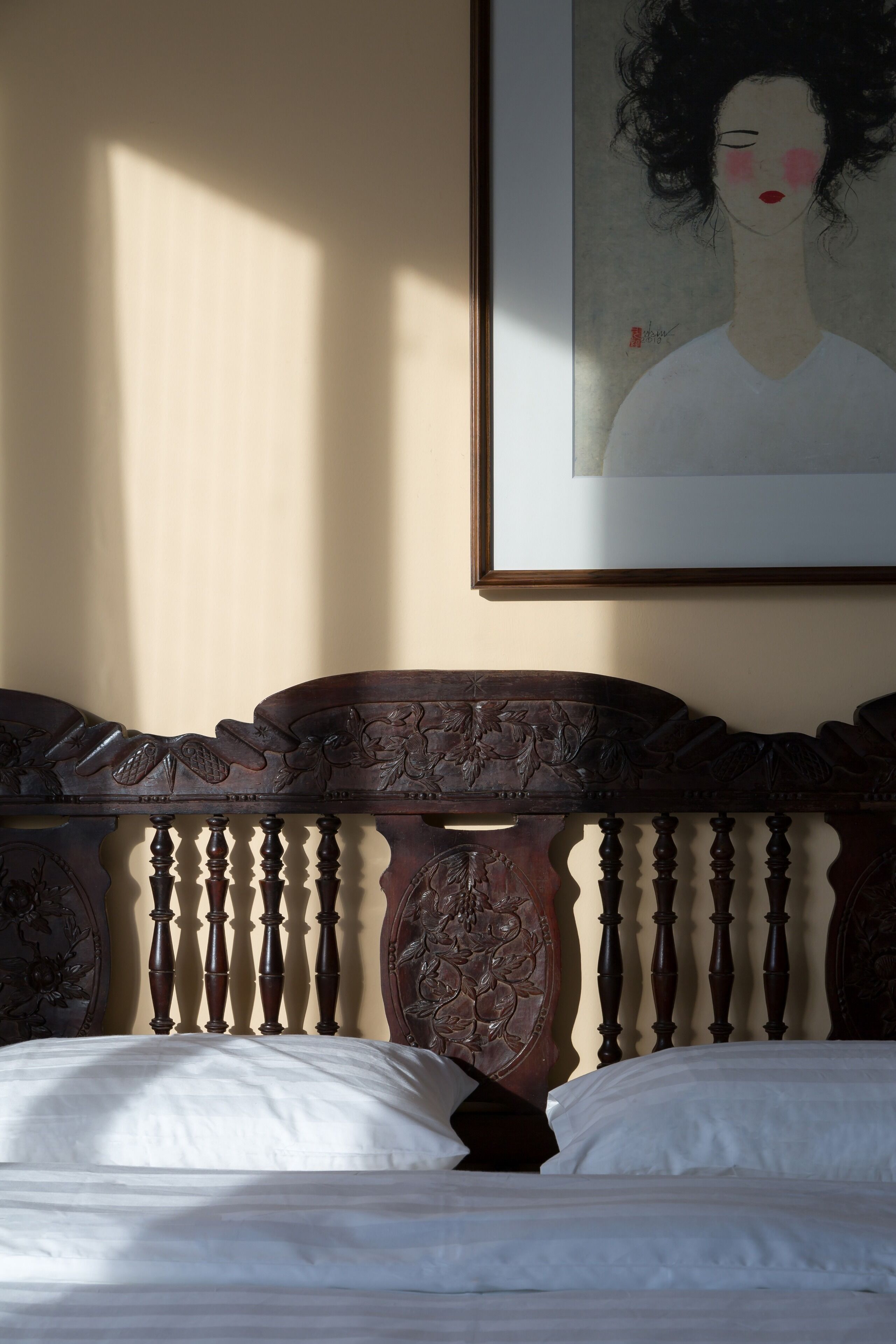 A bed with a carved wooden headboard, a framed artwork above it, and a plain wall.