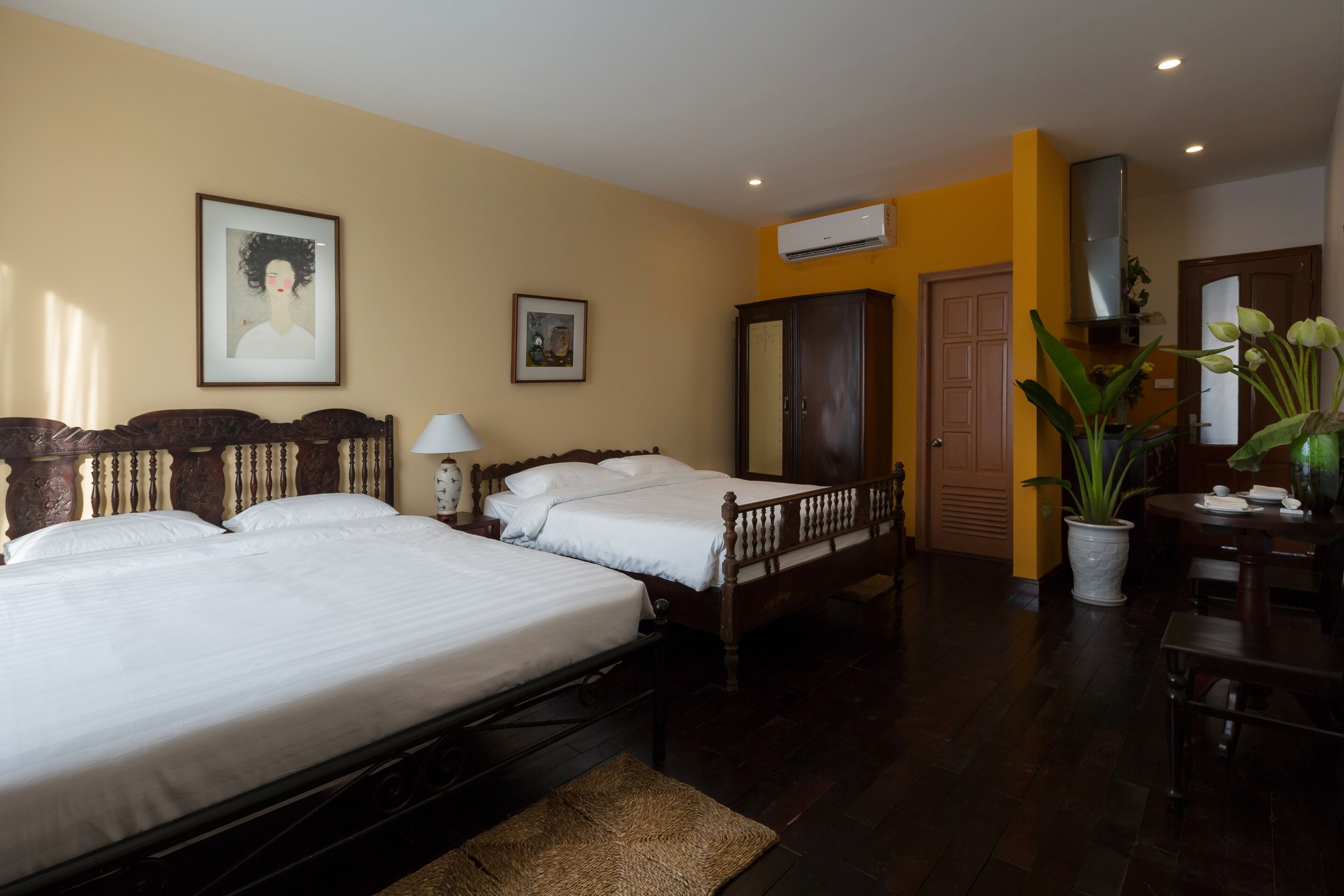 A hotel room with two single beds, a wooden wardrobe, a desk with a chair, a potted plant, and framed artwork on the walls.