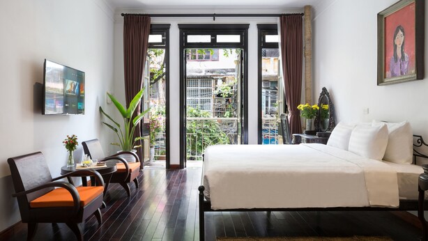 A hotel room with a bed, a desk with a chair, a TV, and a balcony with plants. - Centraltique Downtown Living (Hanoi)
