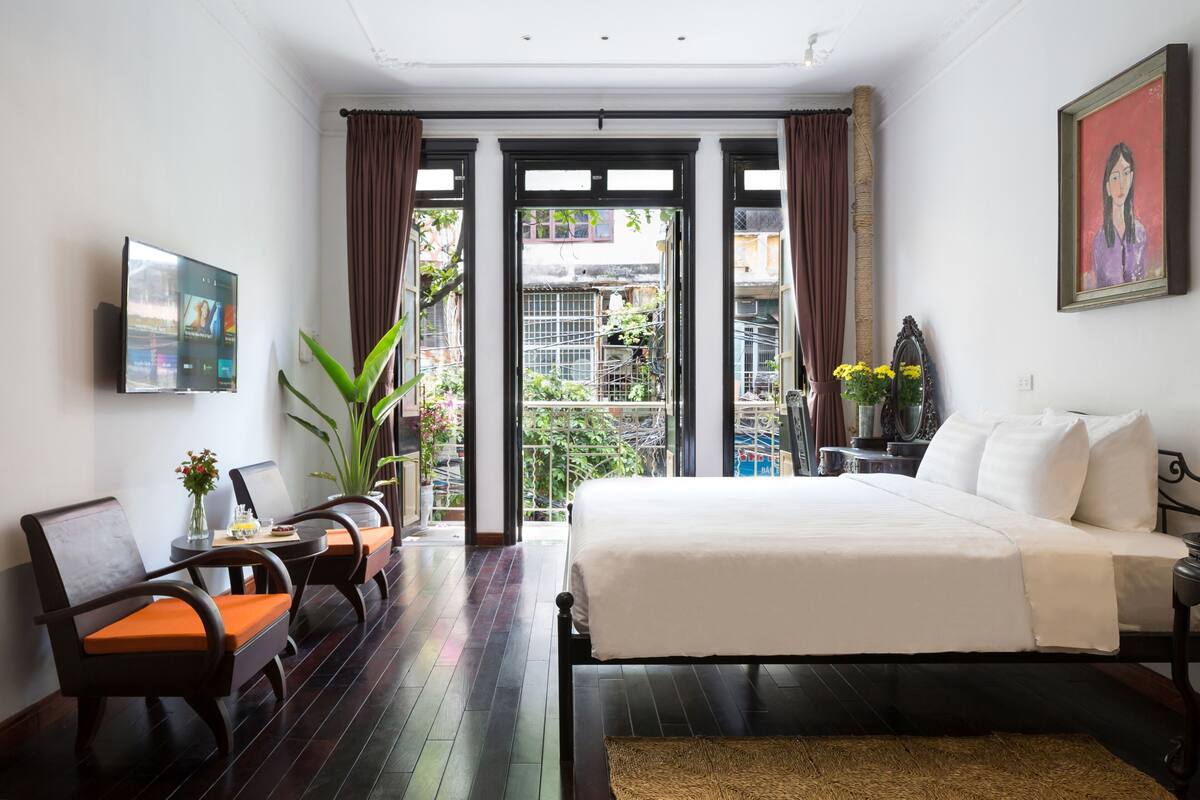 Quarto de hotel com cama, escrivaninha com cadeira, televisão e varanda com plantas.