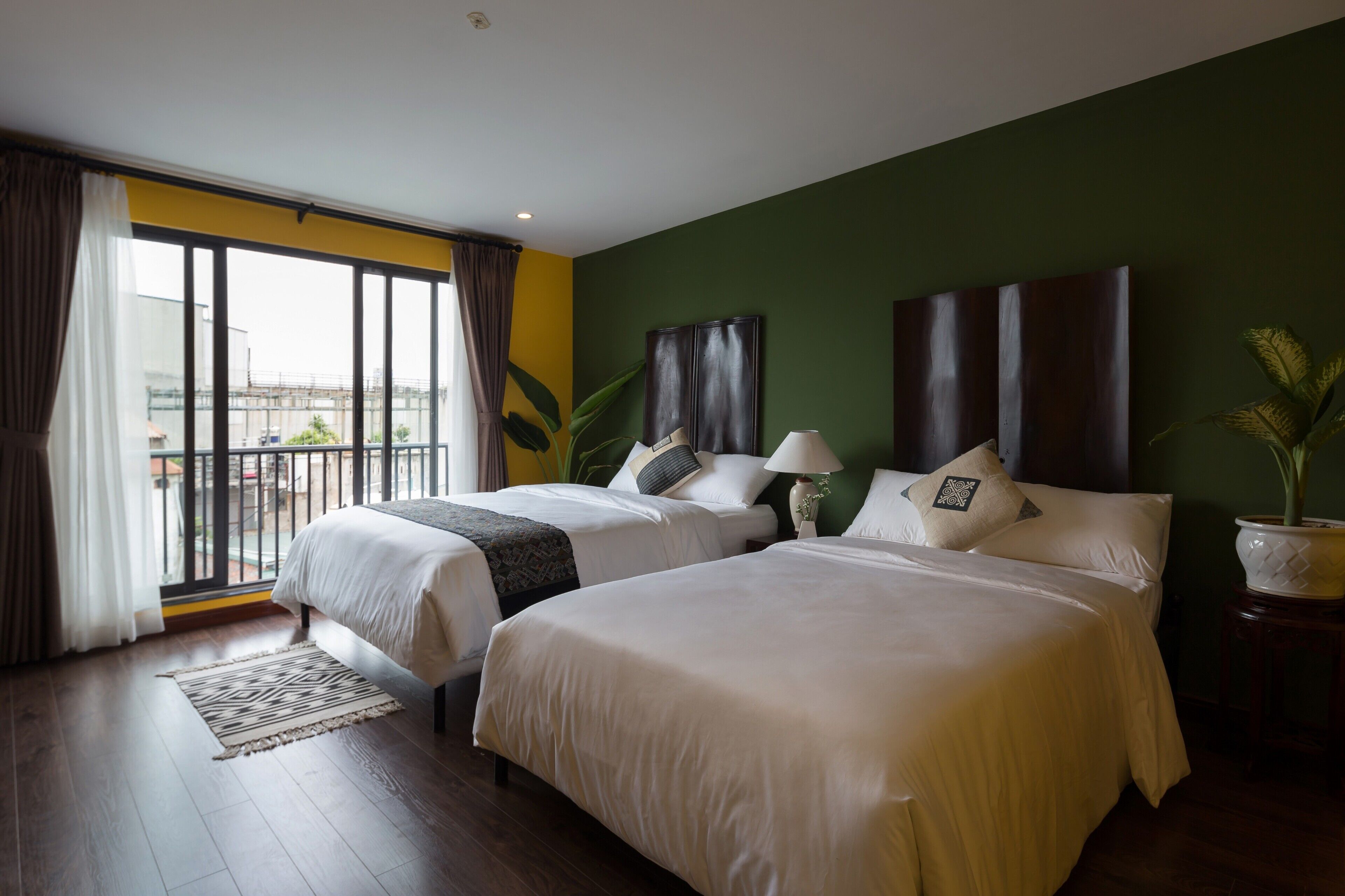 A hotel room with two beds, a large window with curtains, a green wall, and a potted plant.