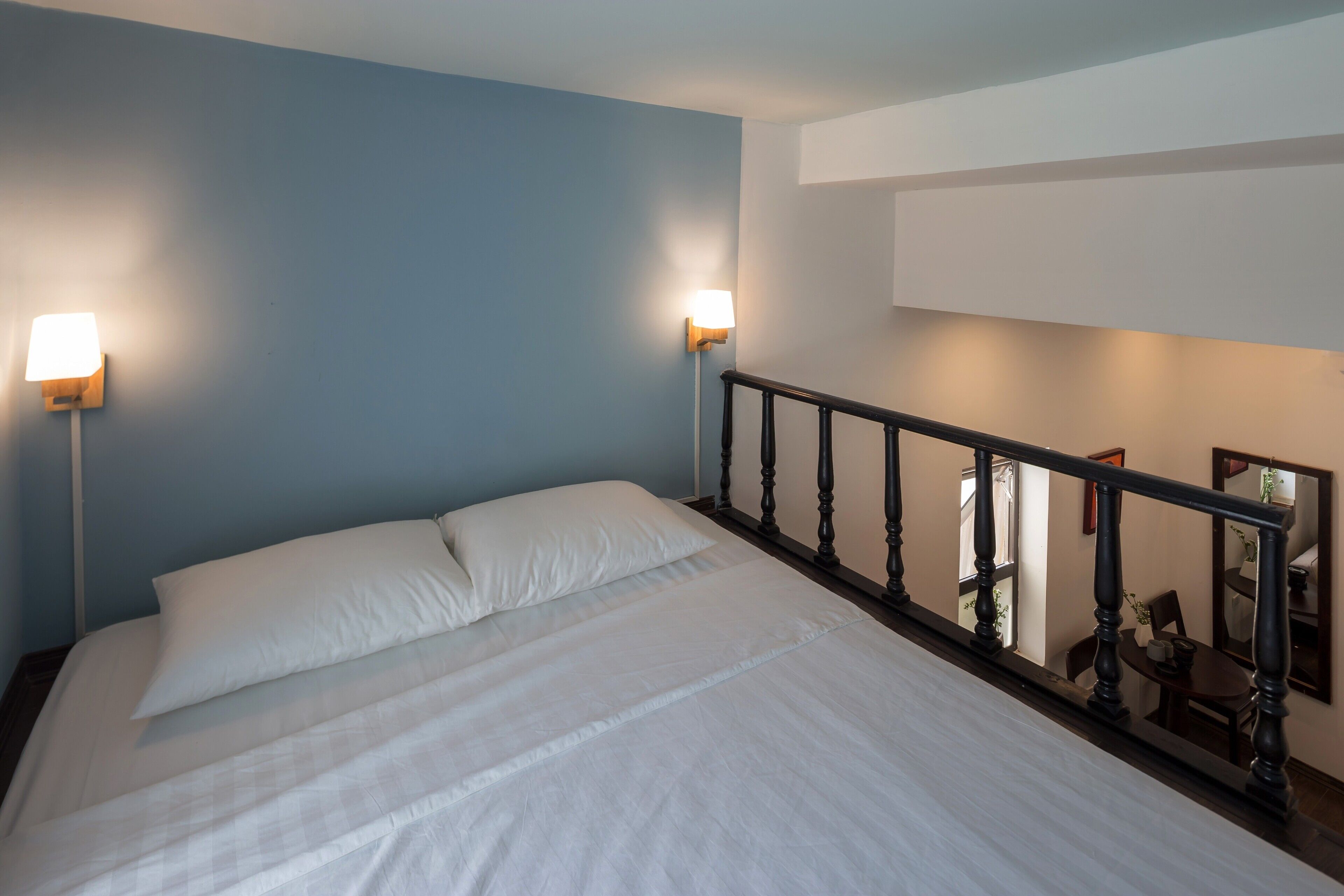 Cama bem arrumada com lençóis brancos, cabeceira de madeira e duas luminárias de leitura fixadas na parede.