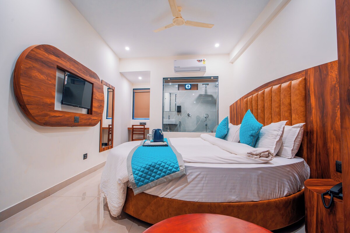 A modern hotel room with a large bed, a wooden headboard, a TV mounted on the wall, and a bathroom with a glass door.