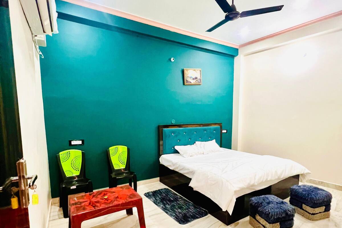 Quarto com paredes verde-água, uma cama, duas cadeiras, um banco, um ventilador de teto e piso de mármore.