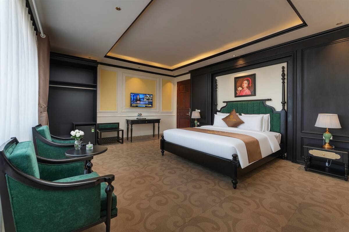 Habitación de hotel con una cama, sillas tapizadas en verde, un escritorio con televisor y una mesita con lámpara.