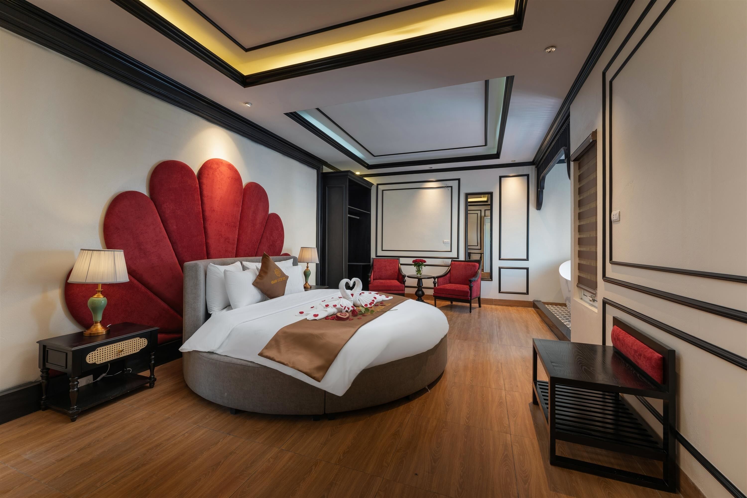 A modern hotel room with a round bed, red accent pillows, a red headboard, and a small table with a lamp.