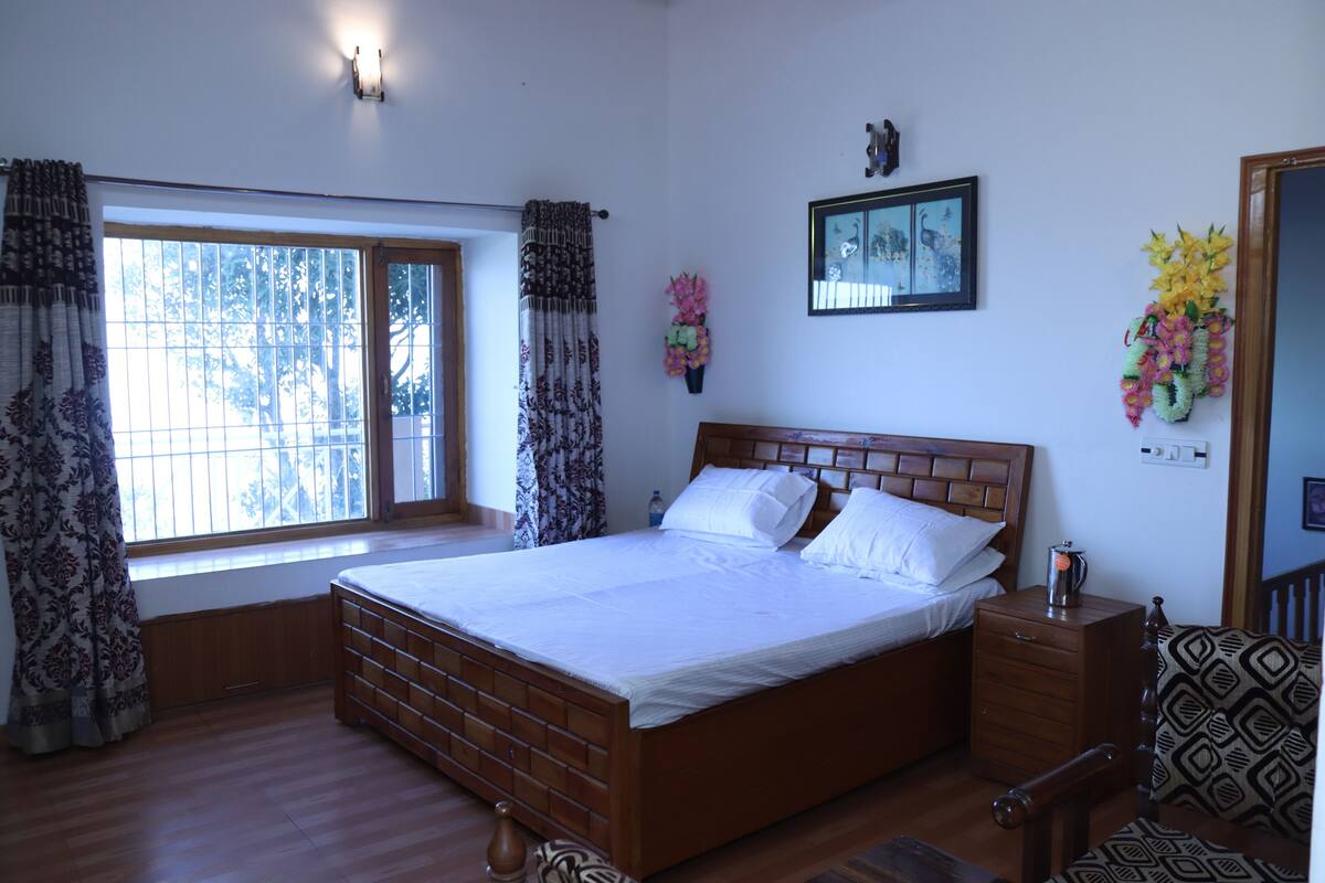 A bedroom with a wooden bed, a nightstand, a chair, a window with curtains, a wall-mounted picture, and a wall-mounted light fixture.