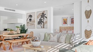 A modern living room with a sofa, coffee table, and dining area. There are framed artworks on the wall and a decorative wall hanging. - Oracle Resort Lvl 22 Beach Views (Broadbeach)