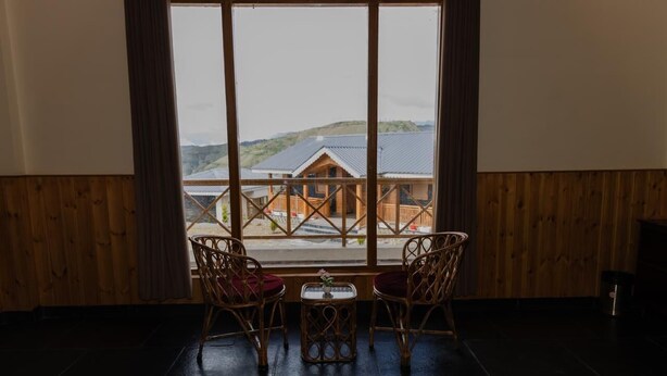 A room with a large window offering a view of a building and hills. There are two wicker chairs and a small table with a vase. - The Meemos Cafe And Cottages (Mussoorie)