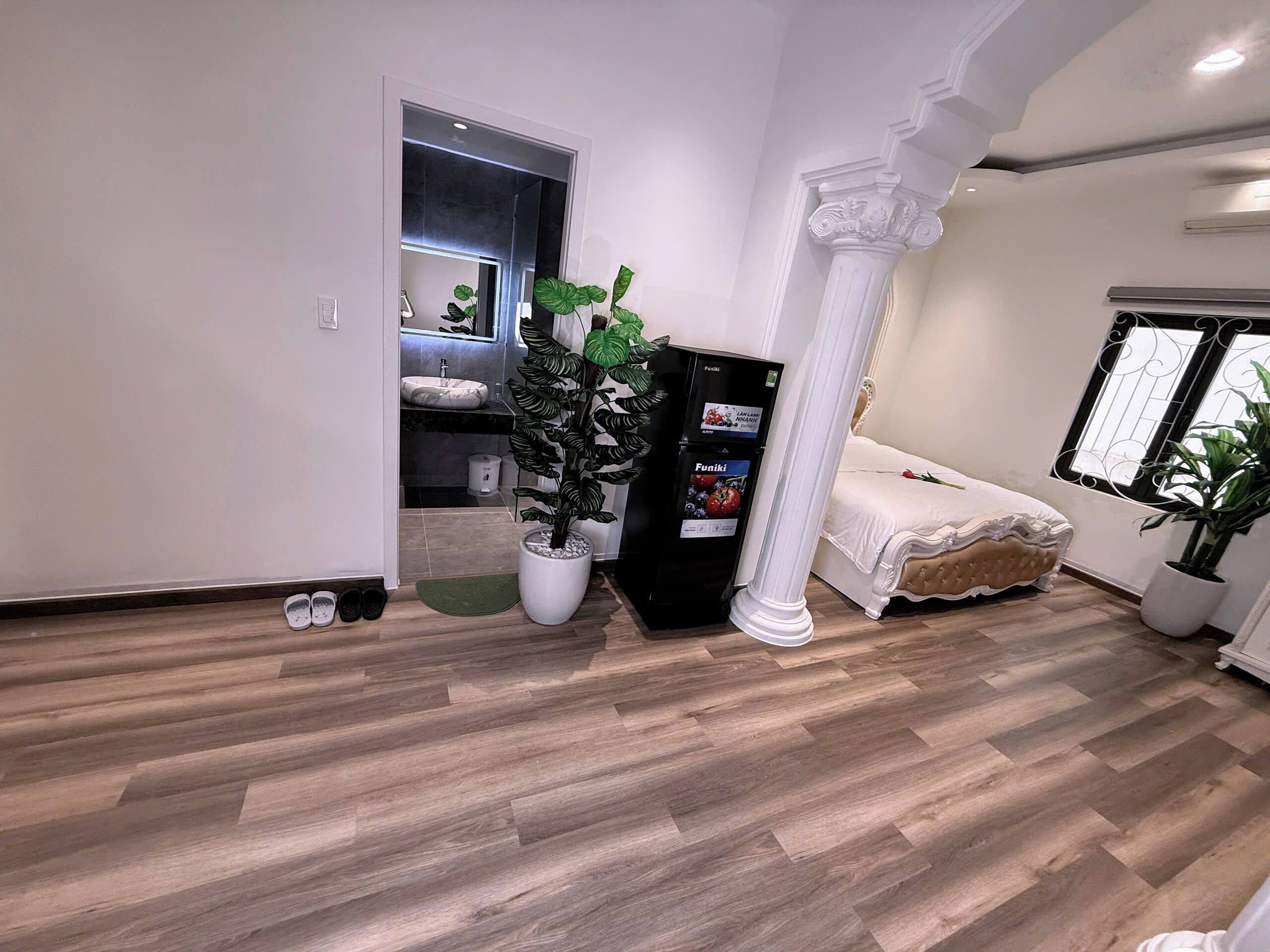 Une chambre moderne avec un lit, une table de chevet, une grande plante en pot, un réfrigérateur noir et une fenêtre ornée de barreaux en fer décoratifs.