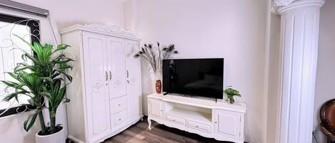 A room with a white cabinet, a television on a white stand, a potted plant, and a wooden floor.