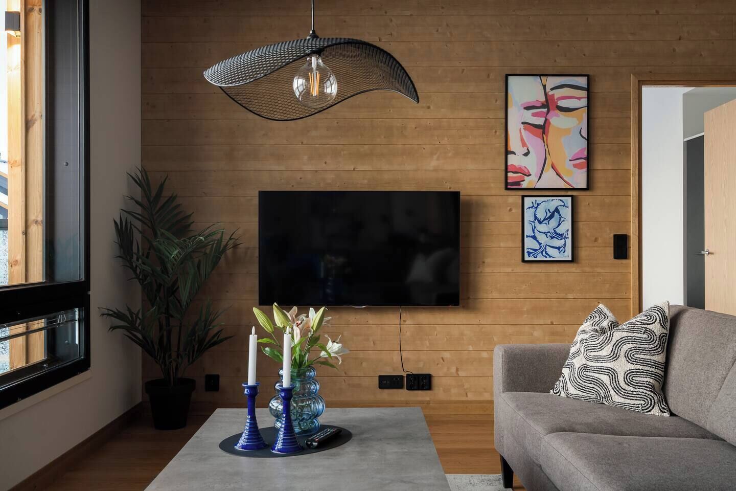 A modern living room with a large flat-screen TV mounted on a wooden wall, a grey sofa with patterned cushions, a coffee table with decorative vases, and a hanging light fixture.