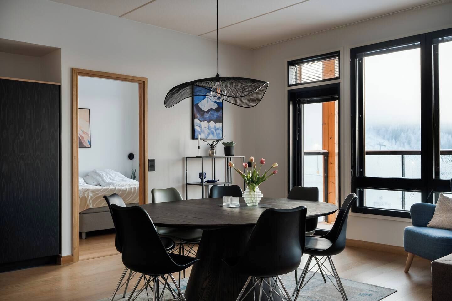 A modern dining area with a round table, black chairs, a hanging light fixture, and a view of the outdoors through large windows.