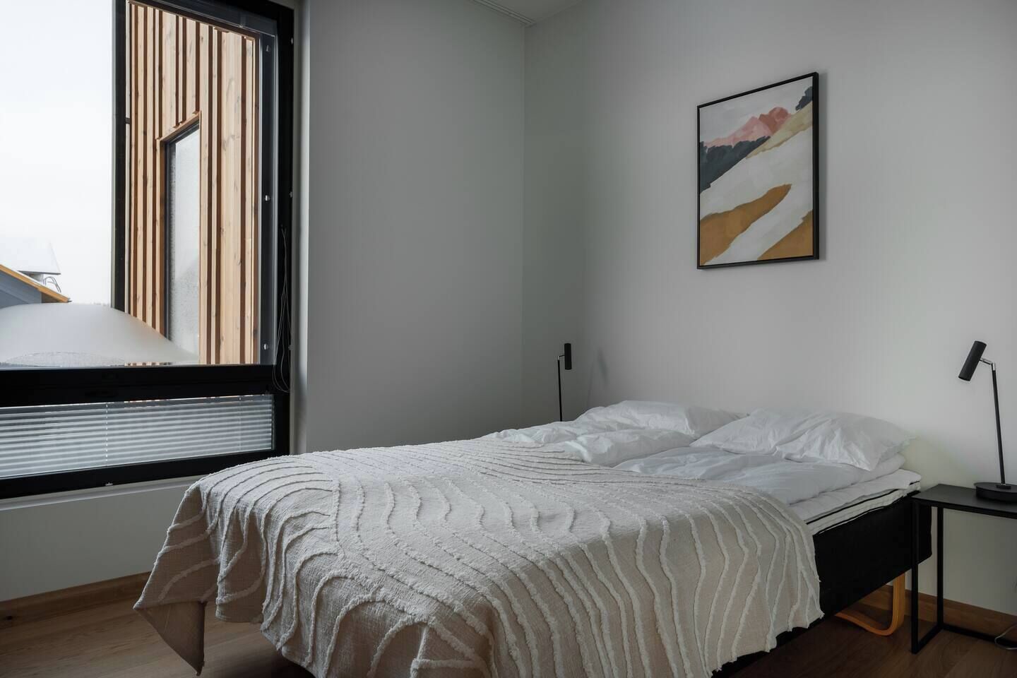 A bedroom with a bed, a bedside table, a lamp, a window with blinds, and a painting on the wall.