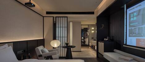 A modern hotel room with a large bed, a seating area, a TV screen displaying hotel services, and a view into the room's interior.
