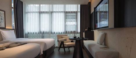 A modern hotel room with two beds, a sofa, a small table, and a large window with curtains.