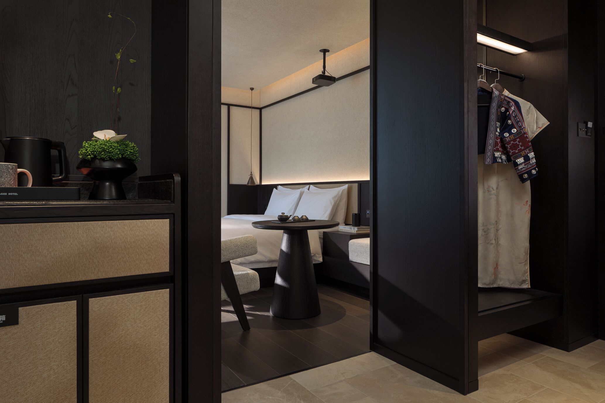 Une chambre d’hôtel moderne avec un décor en bois foncé, une table ronde noire, un lit avec du linge de maison blanc et un portant à vêtements suspendu.