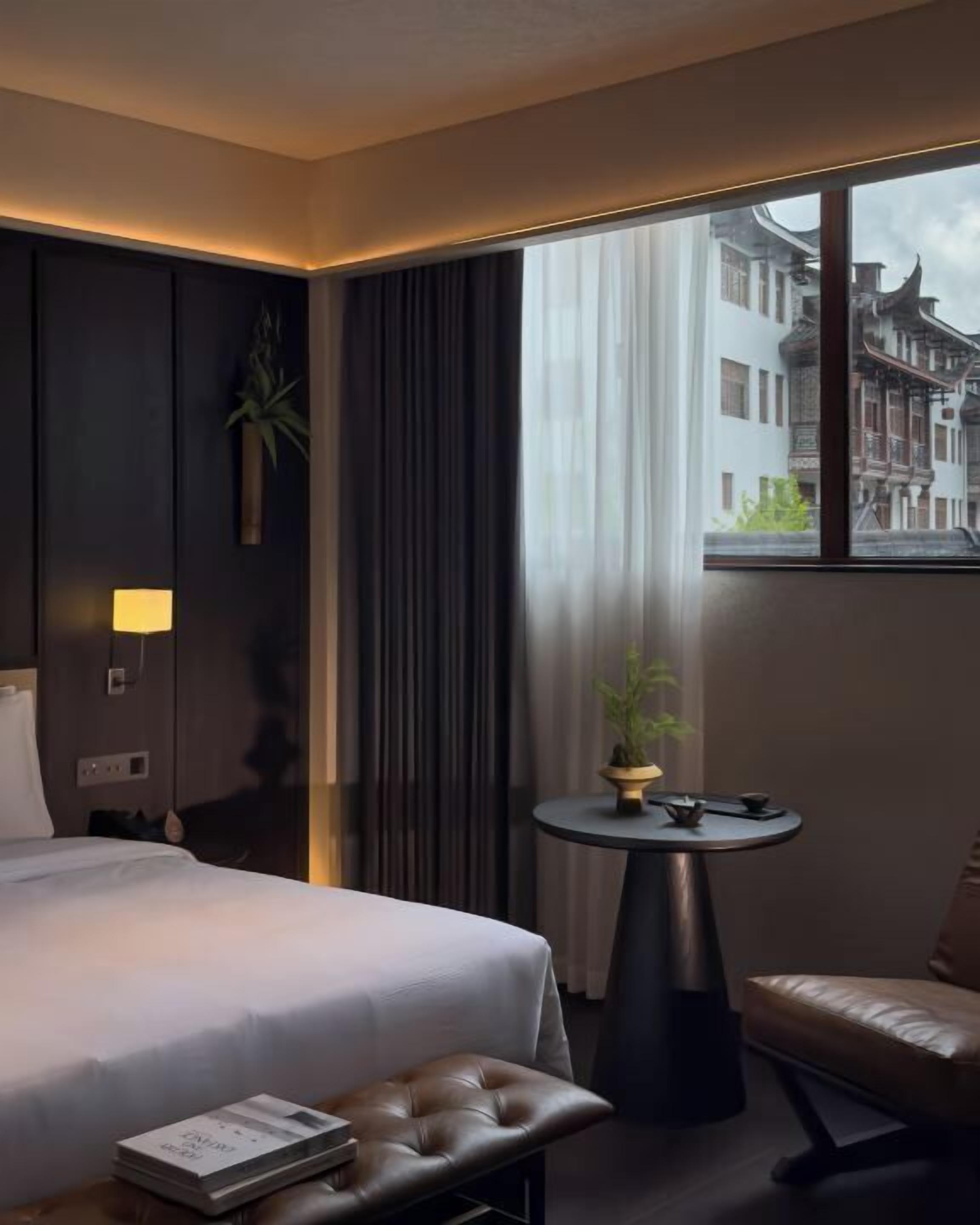 Hotellihuone, jossa on sänky, pieni pyöreä pöytä, jossa on kasvi, tuoli ja ikkunasta avautuva näkymä perinteisiin rakennuksiin.