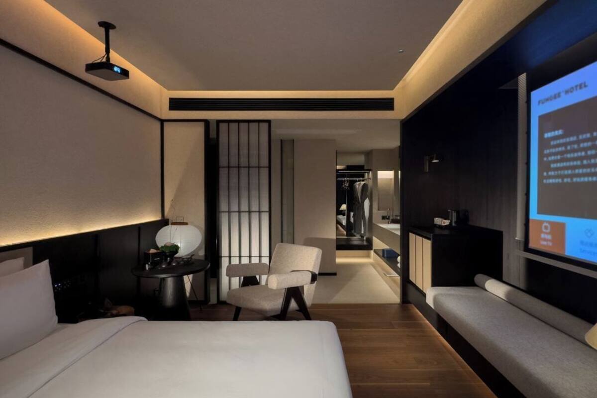 Une chambre d’hôtel moderne équipée d’un lit, d’un coin salon, d’une petite table et d’un écran de télévision affichant des informations sur la chambre.