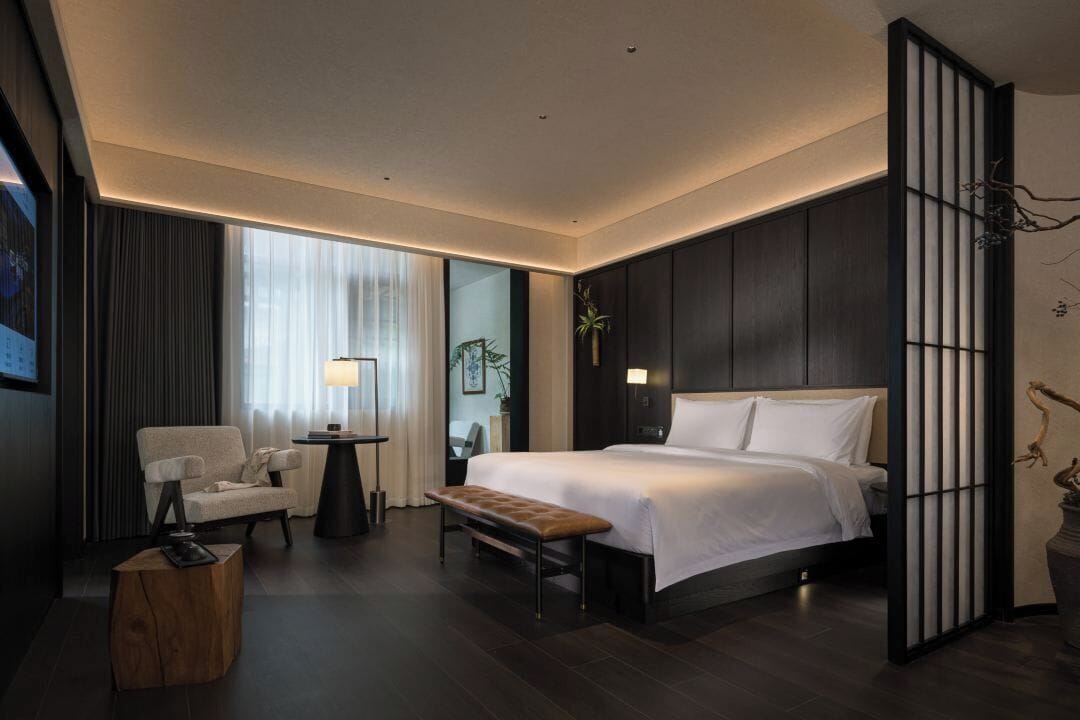 A modern hotel room with a large bed, a sitting area with a chair and a small table, and a large window with curtains.