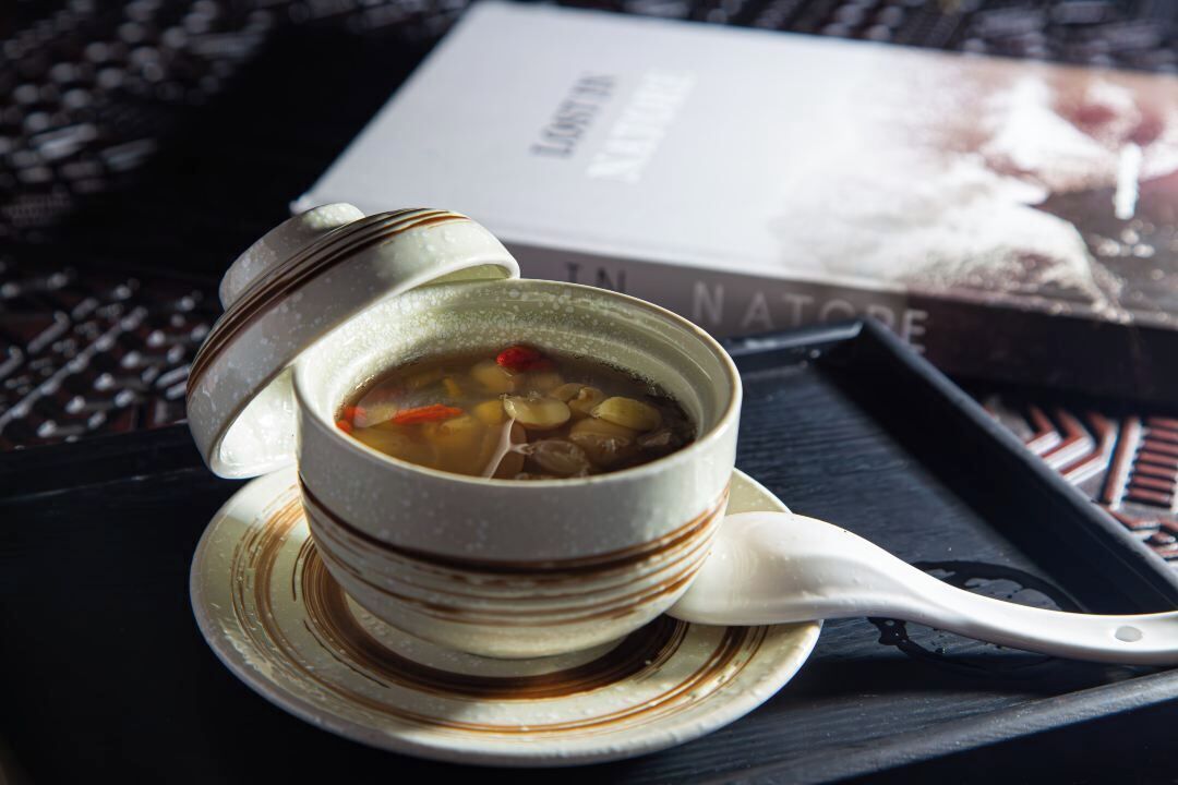 A bowl of soup with a spoon on a saucer, placed on a tray with a book in the background.