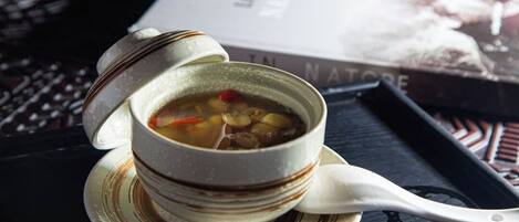 A bowl of soup with a spoon on a saucer, placed on a tray with a book in the background.