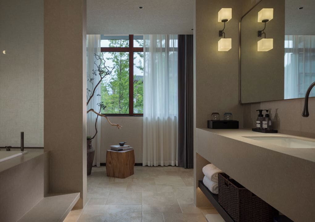 A modern bathroom with a large mirror, a bathtub, and a sink. There is a window with curtains and a view of trees outside.