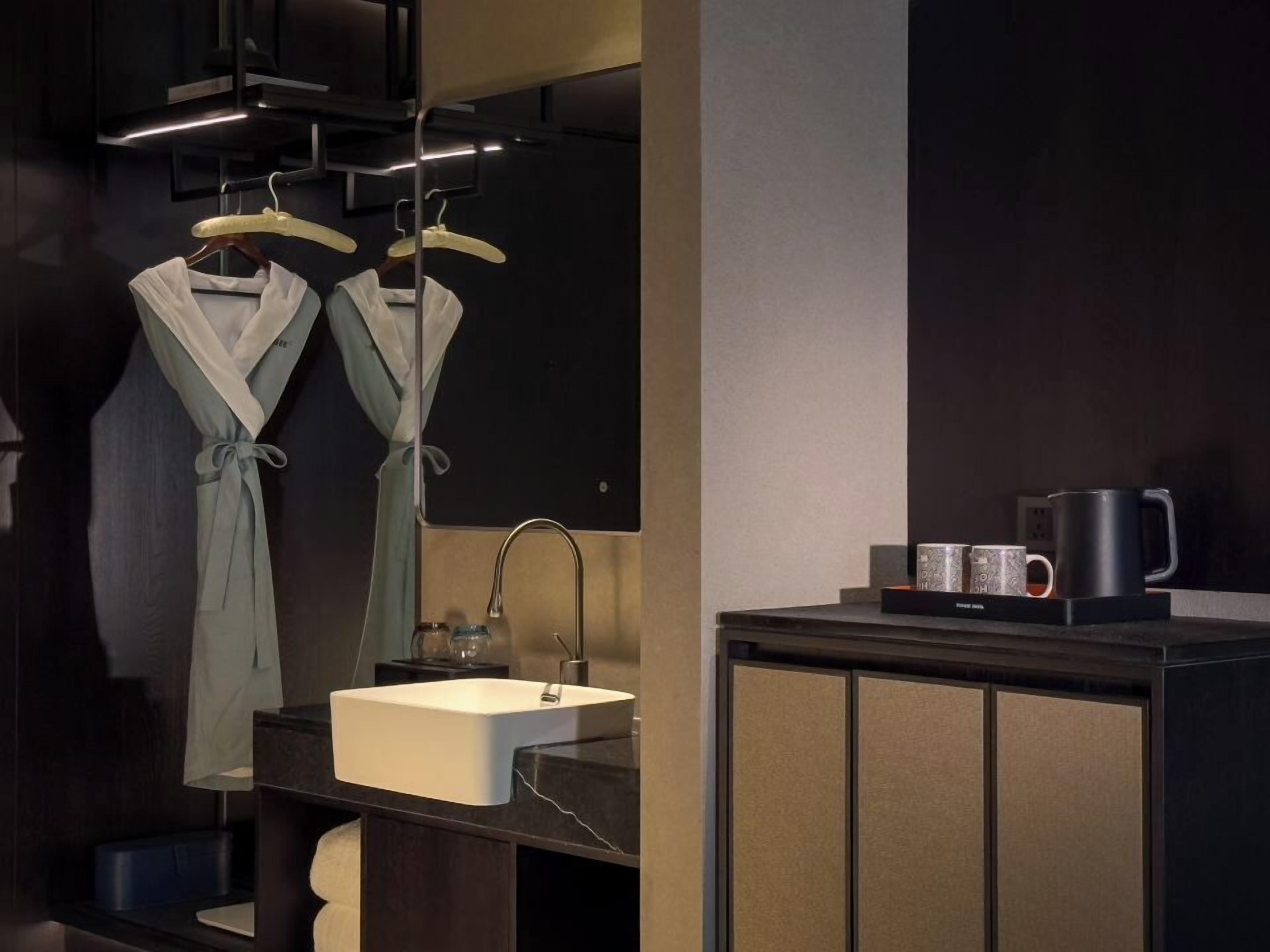 A modern bathroom with a white sink, dark cabinetry, and a mirror above the sink. There are two robes hanging on hooks above the mirror.