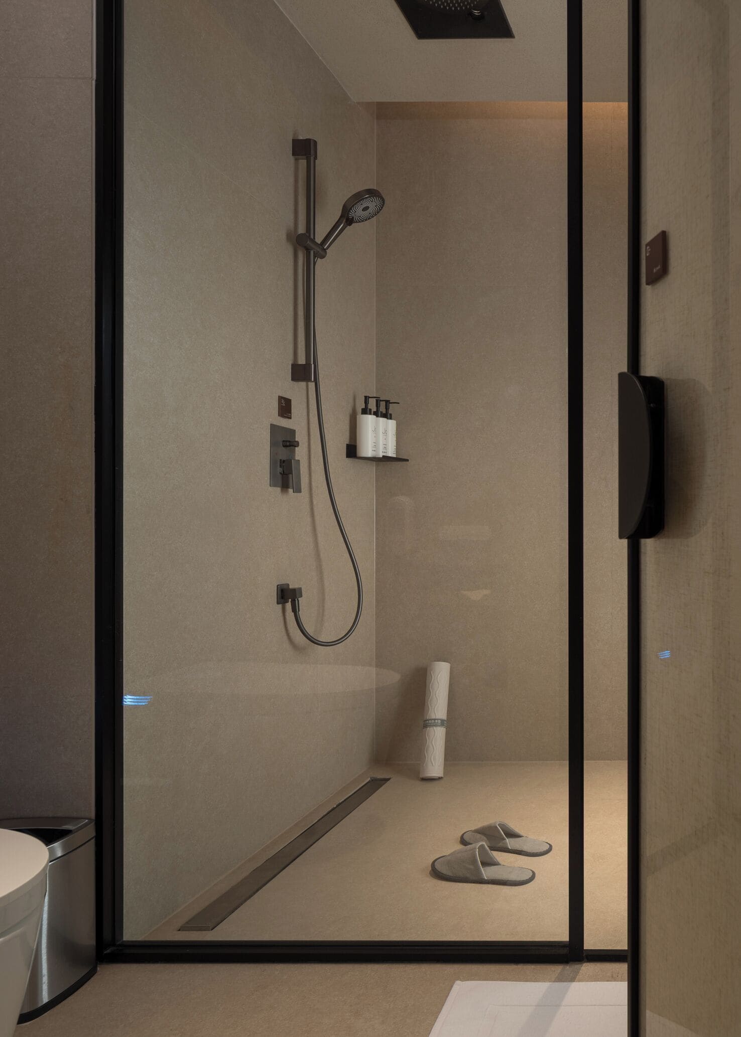 Ein modernes Badezimmer mit Glasabtrennung, einer Dusche inklusive Duschbrausehandstück, einem Handtuch und Hausschuhen auf dem Boden.