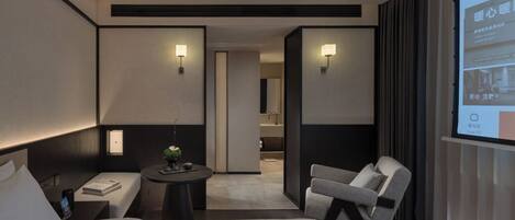 A modern hotel room with a bed, bedside table, chair, and a small desk with a lamp.