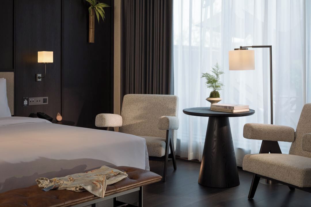 A modern hotel room with a large bed, two armchairs, a small table with a plant, and a floor lamp.