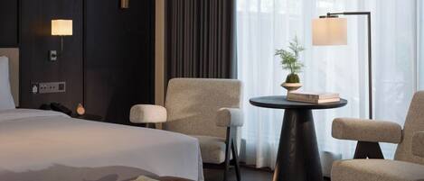 A modern hotel room with a large bed, two armchairs, a small table with a plant, and a floor lamp.