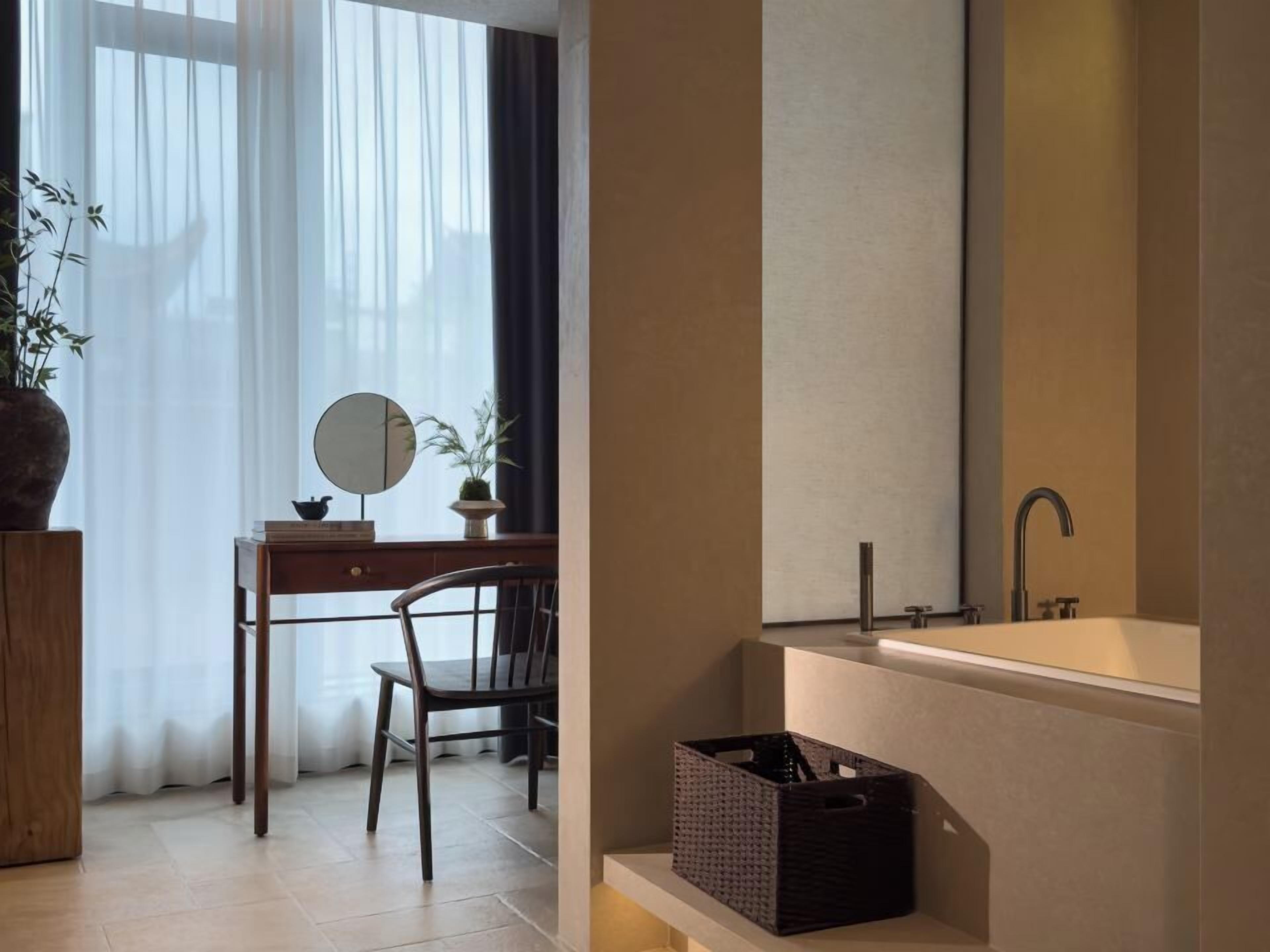 A modern bathroom with a bathtub, a sink, and a mirror. There is a chair and a desk with a plant and a small table.