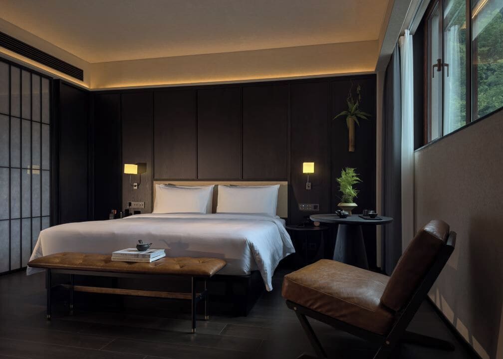 Una habitación de hotel moderna con una cama grande, un banco, una silla, una mesita y una planta en maceta.