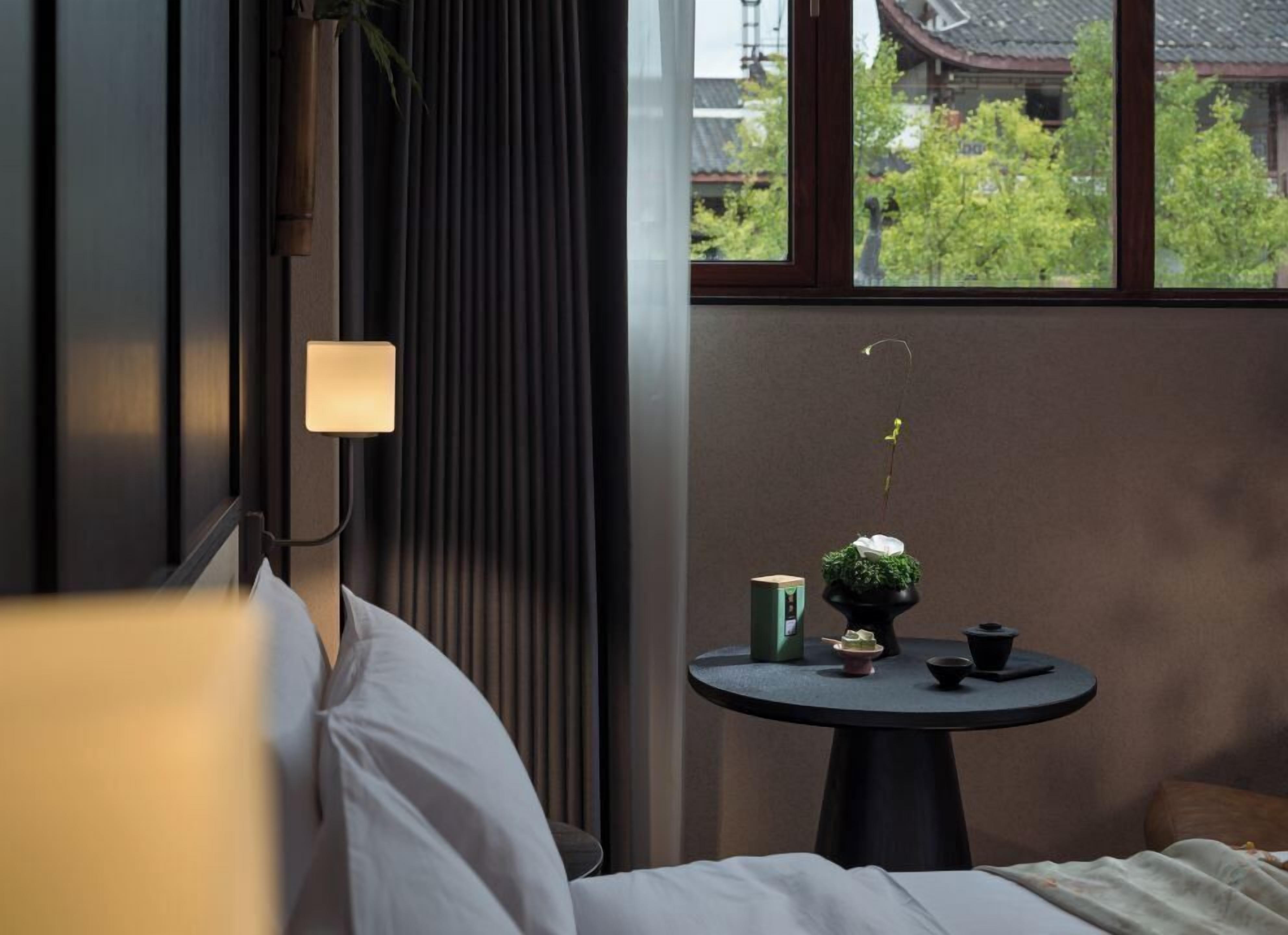 Una habitación de hotel con cama, una mesita de noche con lámpara, una pequeña mesa con una planta y un hervidor, y una ventana con vistas a edificios y árboles.
