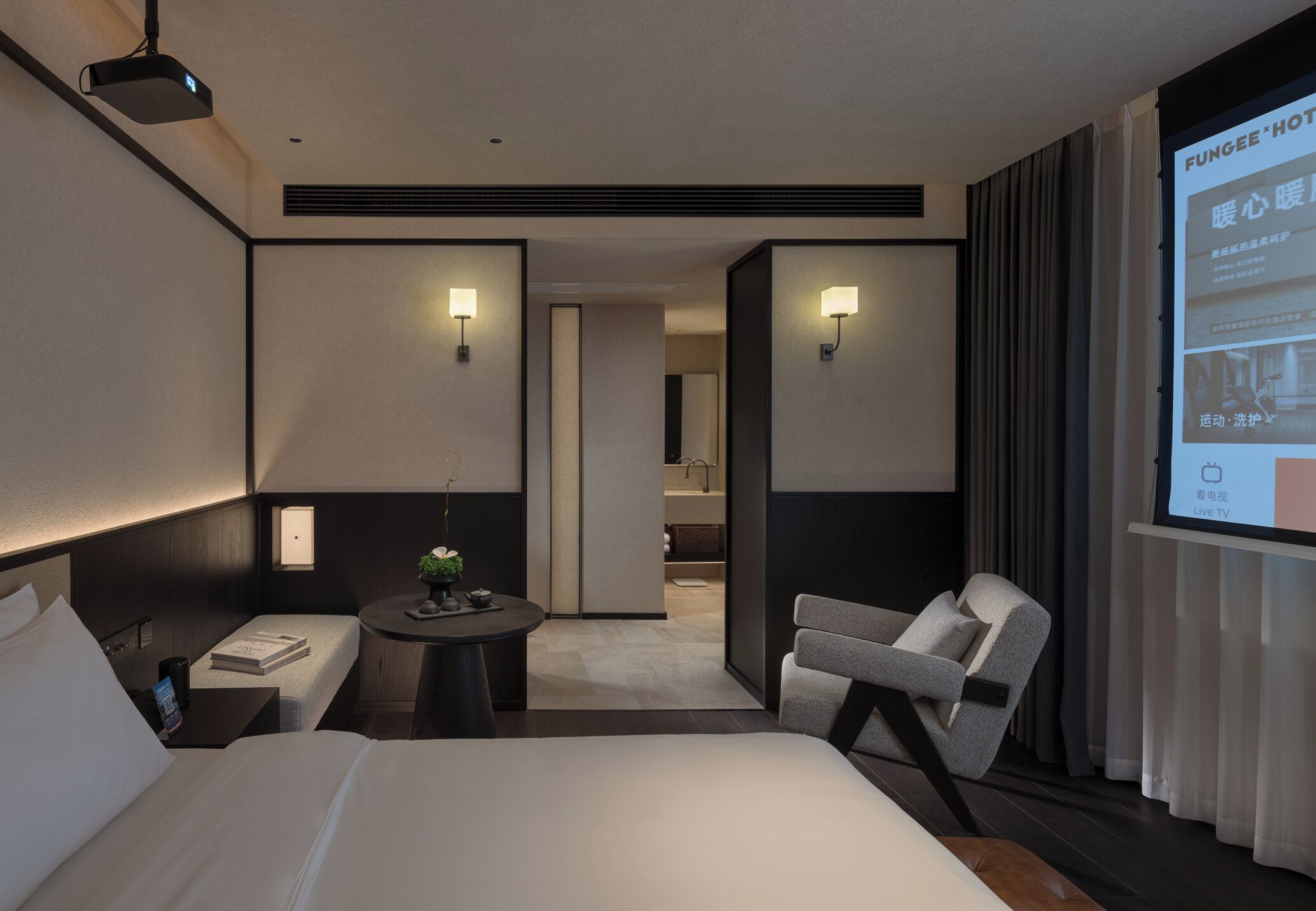 A modern hotel room with a large bed, a desk, a chair, and a TV screen displaying hotel information.