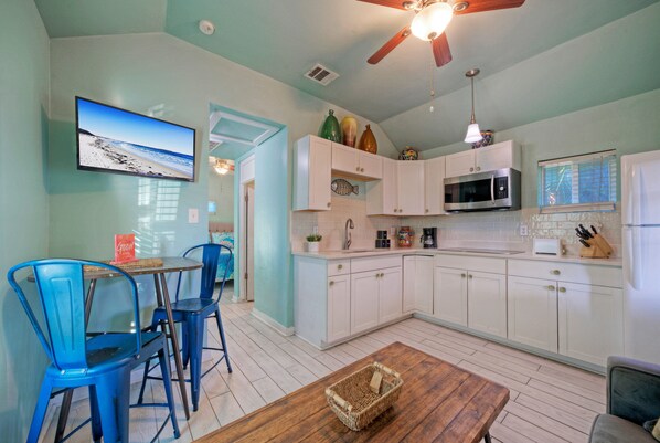 A kitchen with white cabinets, a microwave, and a sink. There is a wooden table with a basket on it. - Cozy Cottage w/ Private Yard in Spanish Village | Casa San Miguel by AvantStay (Port Aransas)