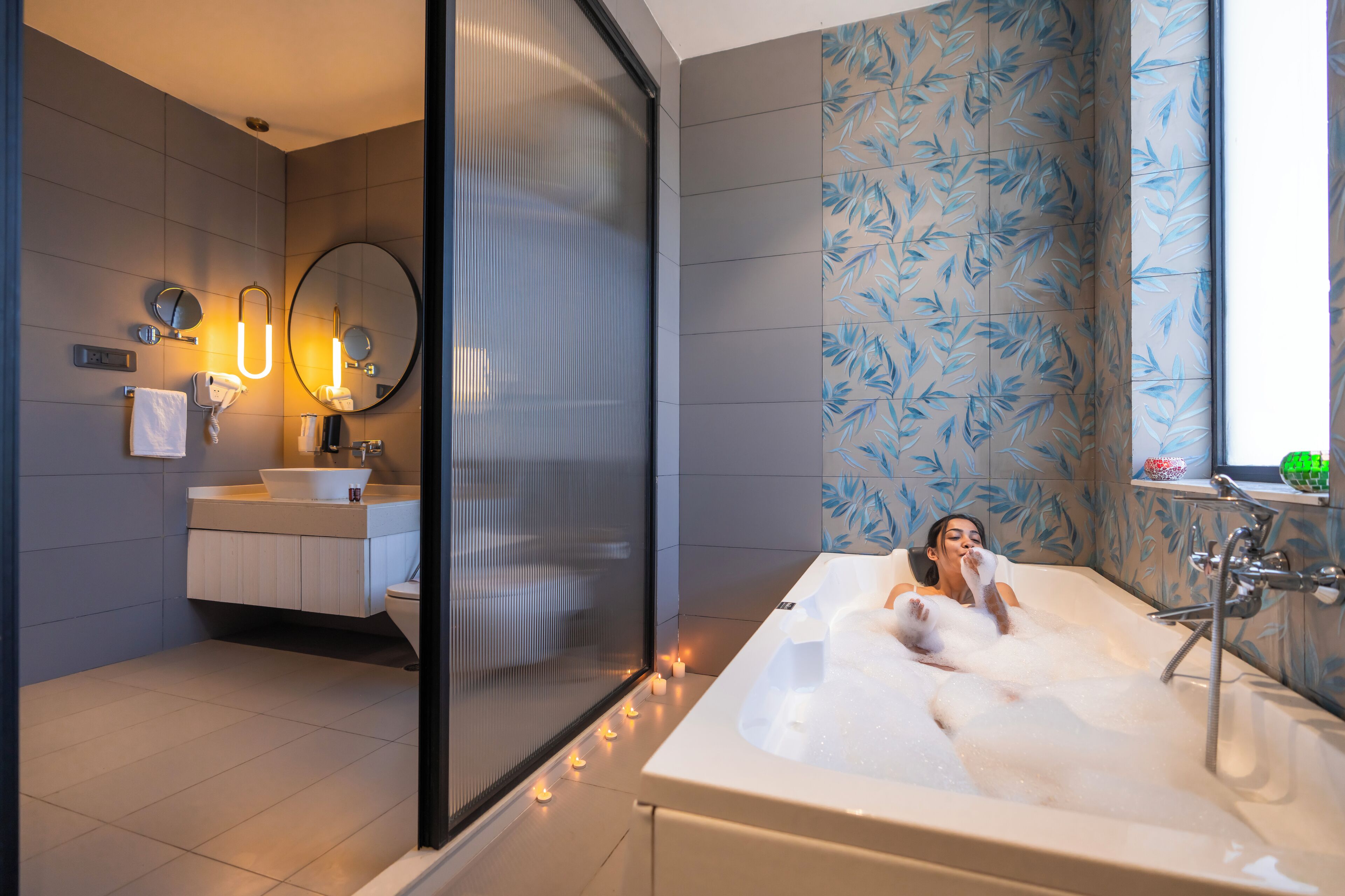 Un baño moderno con una bañera grande, un espejo, un lavabo y un inodoro. Hay velas en el suelo y una ventana con vistas.