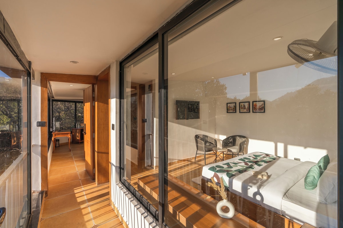 Habitación de hotel moderna con un gran ventanal que da a un balcón, una cama con almohada verde y una zona de comedor con sillas y una mesa.