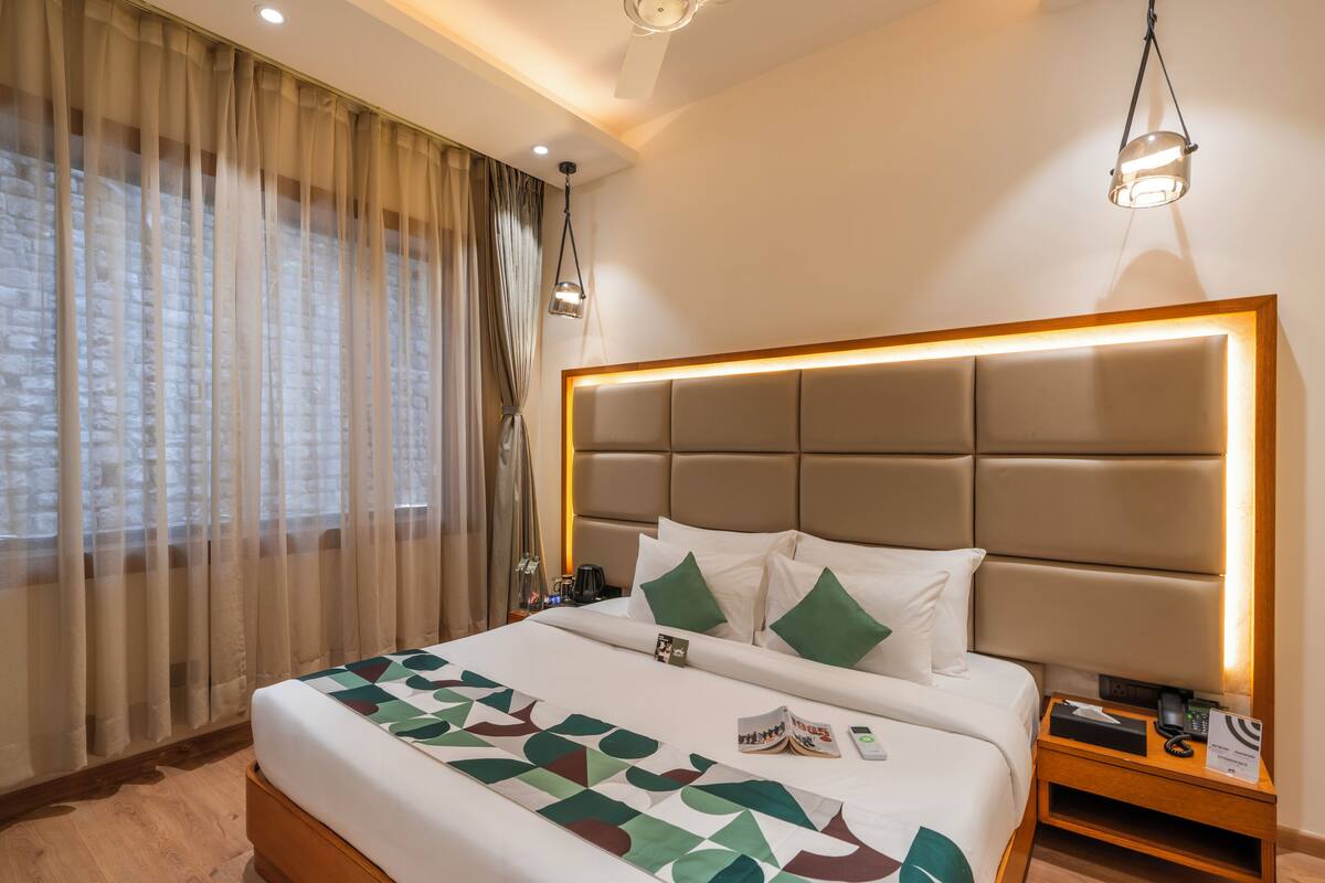 A hotel room with a large bed, a headboard with integrated lighting, a bedside table with a phone and a sign, and a window with sheer curtains.