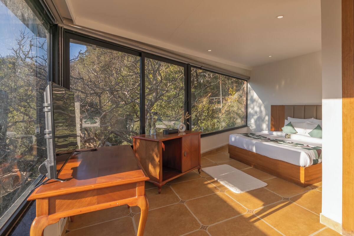 A bedroom with a large window, a wooden desk, a bed, and a nightstand.