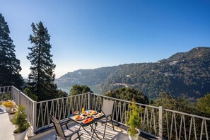 En balkon med et bord dækket til et måltid, med udsigt over et bjerglandskab og et vandområde.