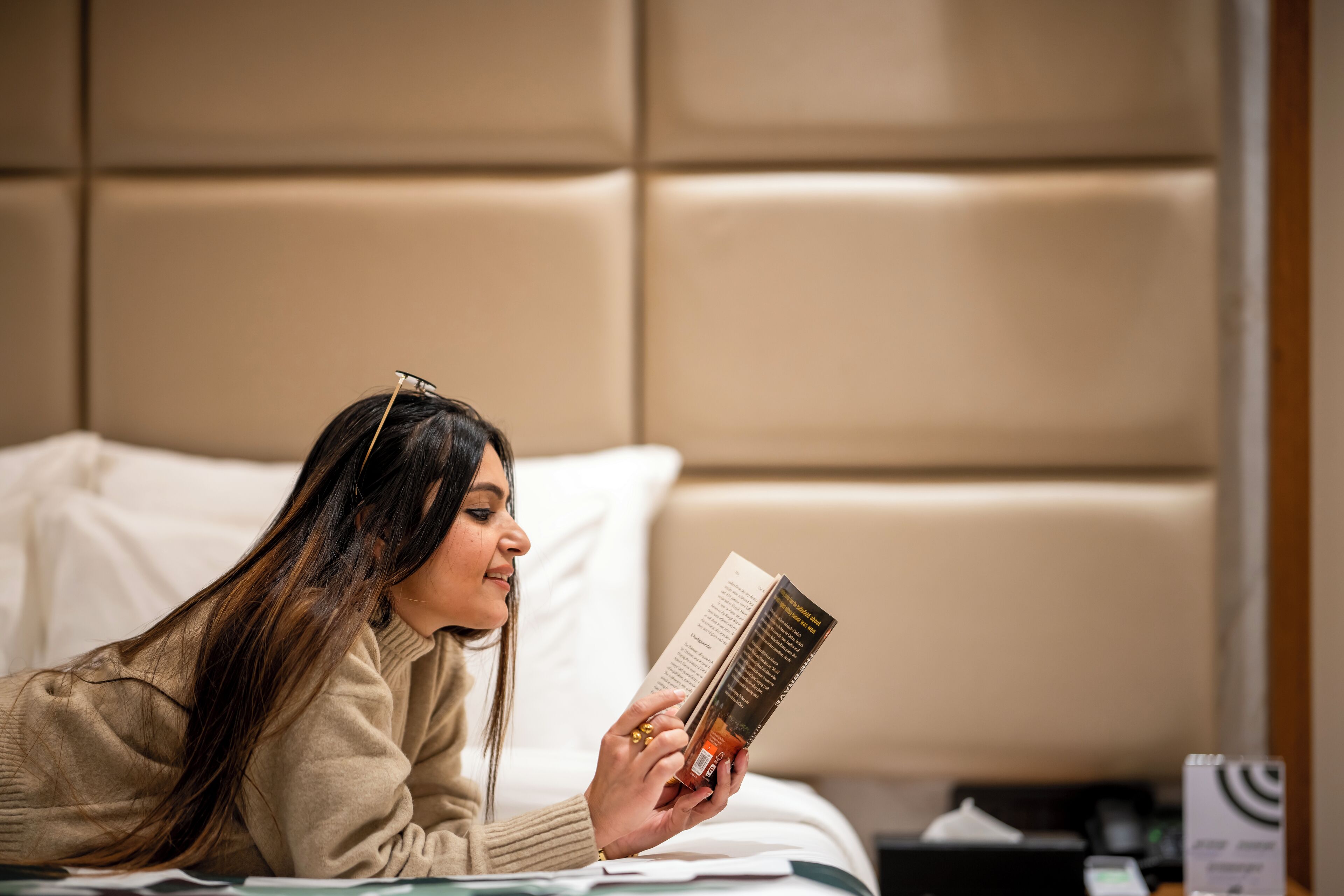 A person lying on a bed with a book, in a room with a padded headboard and white bedding.